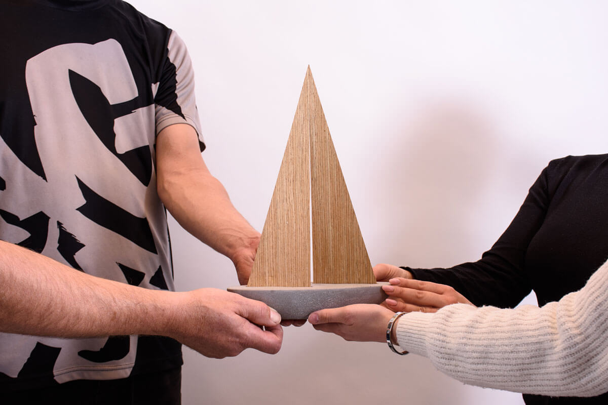 Zwei Personen überreichen ein handgefertigtes Holzobjekt in Segelform als Symbol für das Projekt Hafen zur Arbeitsförderung.